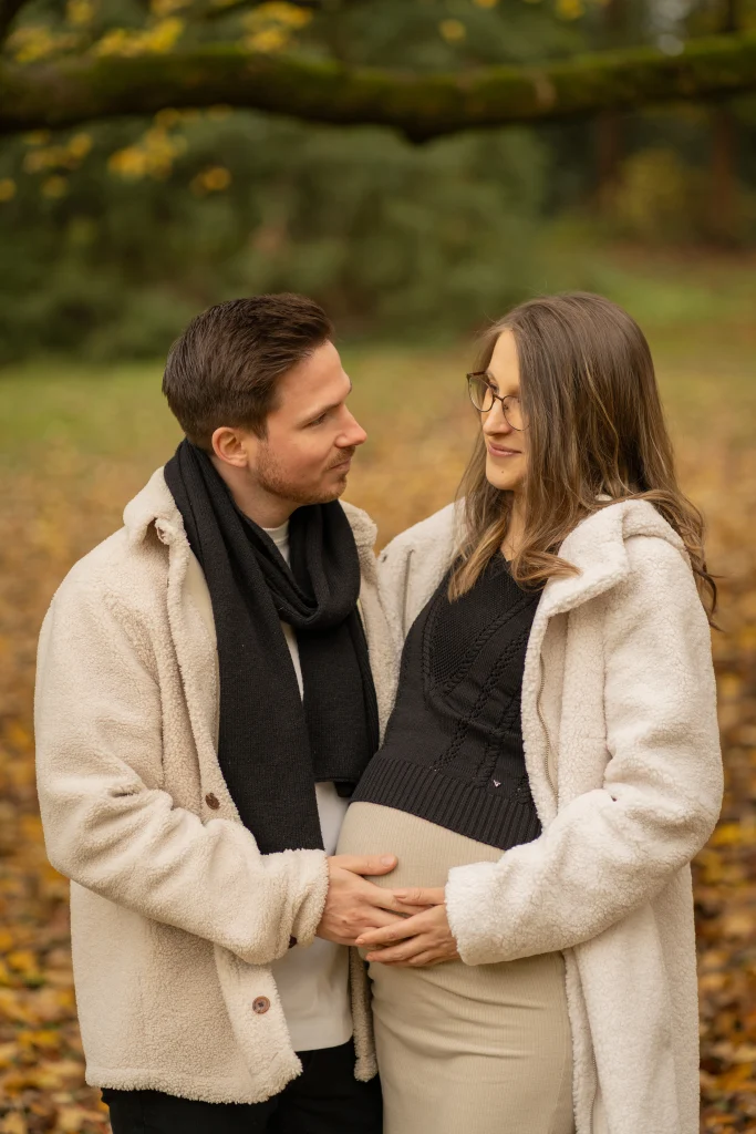 Paar mit Babybauch steht sich lächelnd im Herbstlaub gegenüber – Mosipictures
