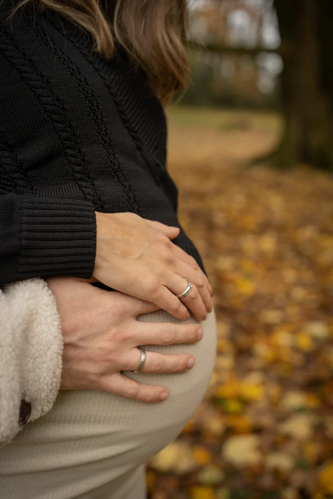 Hände ruhen auf dem Babybauch, Herbstlaub im Hintergrund – Mosipictures