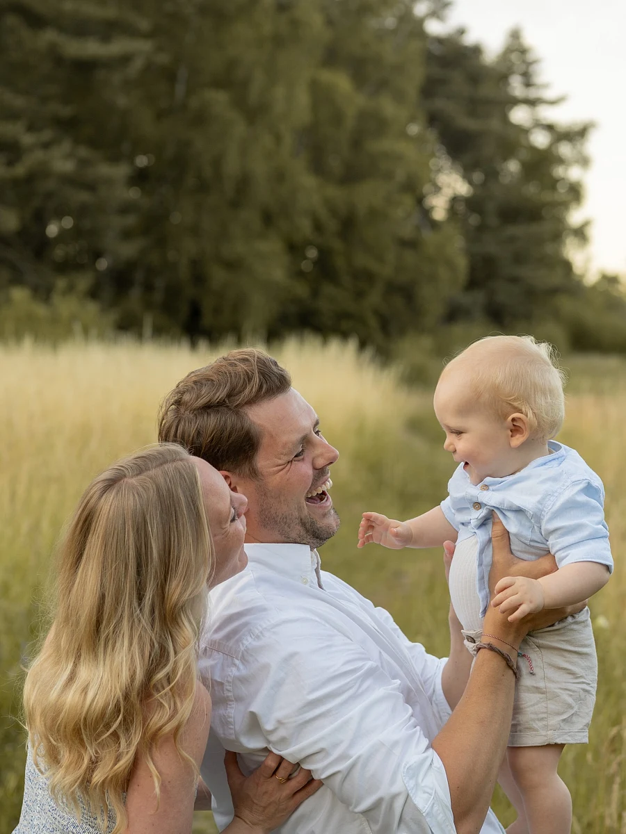 Eltern halten ihr lachendes Kind im hohen Gras im Abendlicht – Familienshooting von Mosipictures